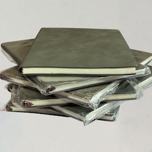 Puede incluir: Una pila de cuadernos verde oliva. Cada cuaderno tiene una cubierta lisa y páginas con bordes blancos. Los cuadernos están envueltos en plástico transparente. La pila está sobre un fondo liso.
