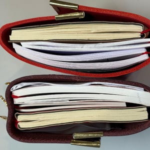 Puede incluir: Dos diarios de cuero, uno rojo y otro burdeos, se muestran abiertos, revelando pilas de papel. Los diarios tienen cierres dorados. Las p&aacute;ginas son una mezcla de rayadas y en blanco, con marcadores de cinta roja.
