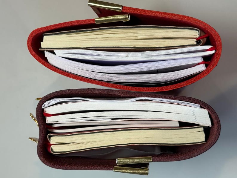 Puede incluir: Dos diarios de cuero, uno rojo y otro burdeos, se muestran abiertos, revelando pilas de papel. Los diarios tienen cierres dorados. Las p&aacute;ginas son una mezcla de rayadas y en blanco, con marcadores de cinta roja.
