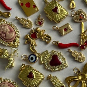 Puede incluir: Una colección de dijes de metal en tono dorado con varios diseños, incluyendo corazones de esmalte rojo, camafeos, manos de hamsa, querubines, un sobre, una carta de juego, un ojo turco y lazos, adornados con cristales claros.
