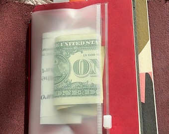 Op de afbeelding: Open leren tijdschrift met een rode binnenkant, met een doorzichtige plastic zak met Amerikaanse valuta. Een klein wit label met het woord "Today" is zichtbaar. Het tijdschrift is open en onthult extra pagina's.