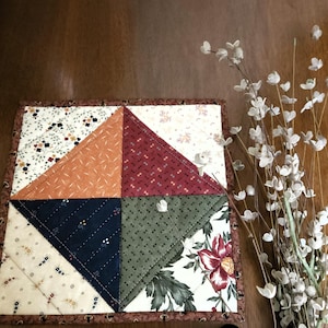 Puede incluir: Un posavasos cuadrado de patchwork con un borde marrón. El posavasos presenta cuatro secciones triangulares en naranja, burdeos, azul marino y verde, cada una con una tela diferente con motivos florales. Un ramo de flores blancas está a la derecha.