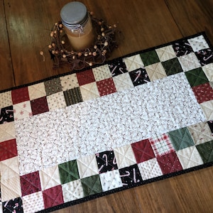 May include: A rectangular quilted table runner featuring a central white panel with a small-scale pattern, surrounded by a border of alternating fabric squares in red, green, black, and cream, some decorated with candy cane and snowflake motifs.