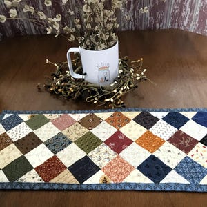 May include: A handmade quilted table runner with a diamond pattern in various colors, including blue, brown, and cream. A white mug with a jar illustration sits on a decorative wreath. The table runner measures approximately 12 inches wide.