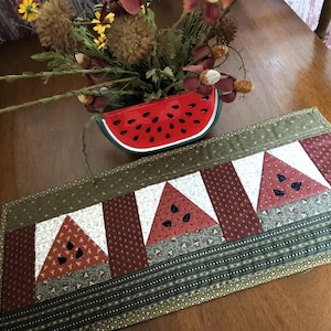 May include: A rectangular quilted table runner featuring a repeating pattern of three red watermelon slice motifs with black seed details, set against white, dark red, and olive green fabric panels with small floral and geometric prints.
