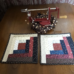 May include: Two square quilted placemats with a log cabin design in shades of cream, red, blue, and black. A vintage red apple peeler sits atop a decorative wreath. The placemats are on a wooden table.