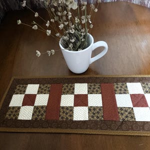 May include: A rectangular brown quilted table runner with a patchwork design. The runner features alternating squares of white, brown, and patterned fabric, with a solid brown stripe in the center. A white mug with dried flowers sits on the table.