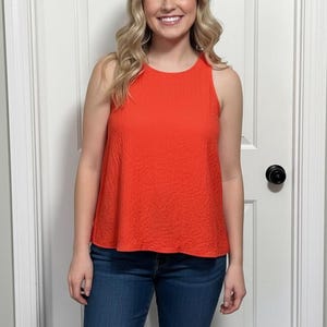Può includere: Una donna con un top arancione brillante senza maniche e jeans blu è in piedi davanti a una porta bianca. Il top ha un aspetto strutturato. La donna ha lunghi capelli biondi ondulati e sorride.
