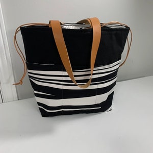 Peut inclure: Sac fourre-tout à rayures noires et blanches avec des anses en cuir marron. Le sac a une fermeture éclair et une grande poche sur le devant.