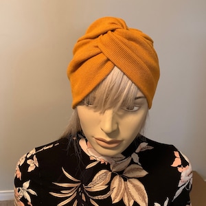 Könnte beinhalten: Ein turbanartiger Hut in Senfgelb aus weichem, geripptem Stoff. Der Hut wird von einer Mannequin mit einem schwarz-weiß gemusterten Hemd getragen.