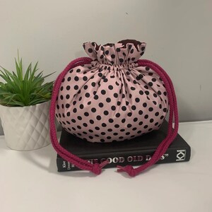 Puede incluir: Una bolsa de cord&oacute;n de lunares rosa y negro con un borde superior con volantes y cordones rosas. La bolsa est&aacute; encima de un libro.