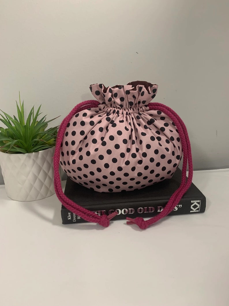 Puede incluir: Una bolsa de cord&oacute;n de lunares rosa y negro con un borde superior con volantes y cordones rosas. La bolsa est&aacute; encima de un libro.