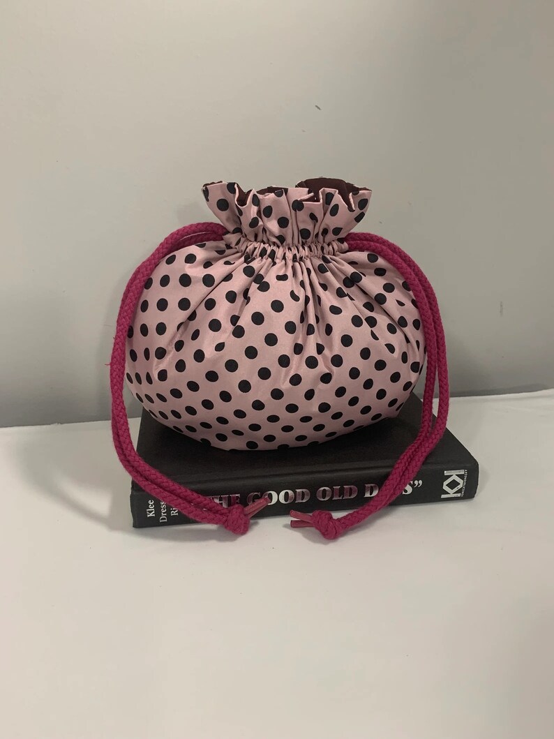 Puede incluir: Bolsa de cord&oacute;n rosa con lunares negros. La bolsa est&aacute; encima de un libro titulado "The Good Old Days".