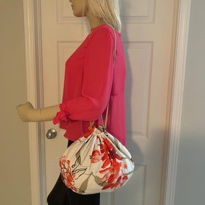 Puede incluir: Una bolsa blanca de cord&oacute;n con estampado floral con flores rojas y rosas. La bolsa tiene una correa larga marr&oacute;n y la sostiene un maniqu&iacute;.