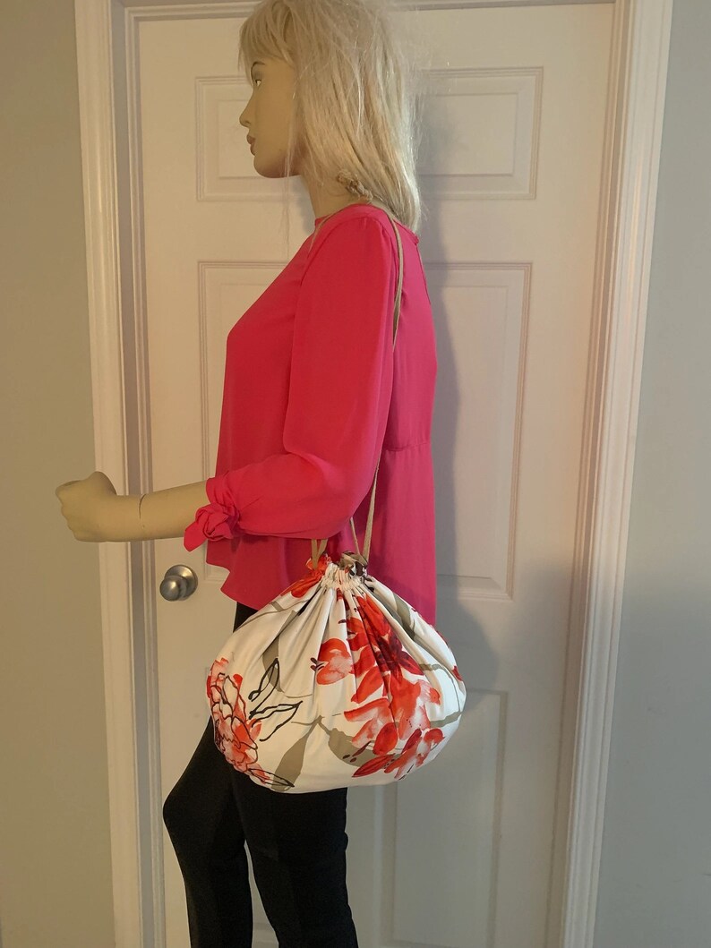 Puede incluir: Una bolsa blanca de cord&oacute;n con estampado floral con flores rojas y rosas. La bolsa tiene una correa larga marr&oacute;n y la sostiene un maniqu&iacute;.