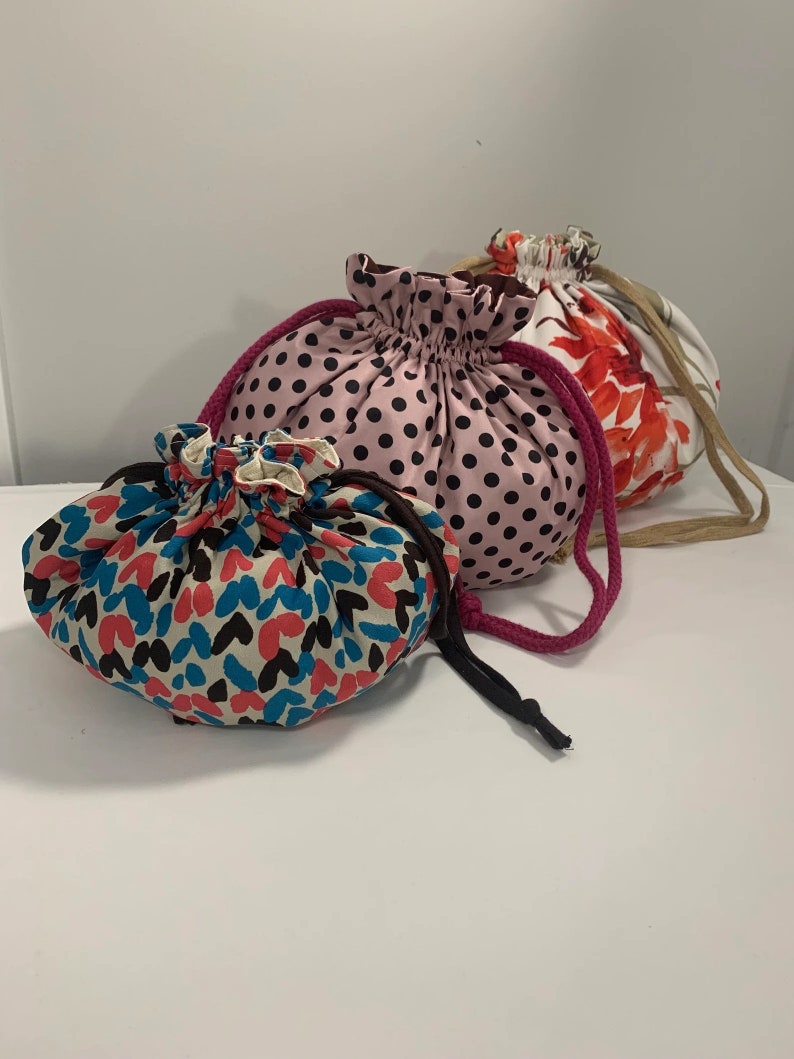 Puede incluir: Tres bolsas de cord&oacute;n hechas de tela. La bolsa m&aacute;s peque&ntilde;a es azul y blanca con un patr&oacute;n de coraz&oacute;n. La bolsa del medio es rosa y negra con un patr&oacute;n de lunares. La bolsa m&aacute;s grande es blanca con un patr&oacute;n floral rojo.