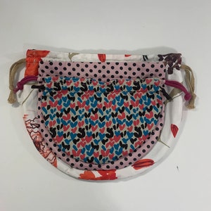 Puede incluir: Tres bolsas de tela con cord&oacute;n de ajuste y diferentes patrones. La bolsa superior es blanca con flores rojas, la bolsa del medio es rosa con lunares negros y la bolsa inferior es beige con un patr&oacute;n de corazones rojos, azules y negros.