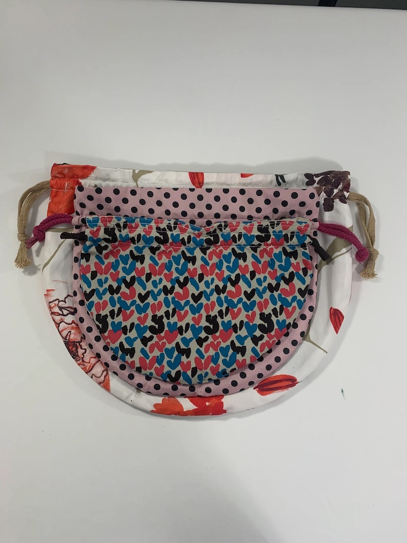 Puede incluir: Tres bolsas de tela con cord&oacute;n de ajuste y diferentes patrones. La bolsa superior es blanca con flores rojas, la bolsa del medio es rosa con lunares negros y la bolsa inferior es beige con un patr&oacute;n de corazones rojos, azules y negros.