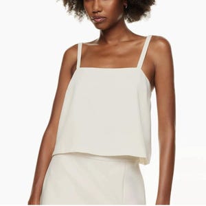 May include: Cream-colored square-neck top with wide shoulder straps. The top is paired with a matching skirt, creating a coordinated outfit. The fabric appears smooth and has a slight sheen, suggesting a formal or semi-formal style.