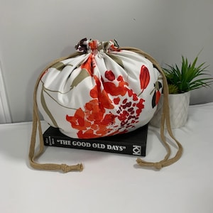 Puede incluir: Una bolsa de cord&oacute;n blanco con estampado floral en tonos naranja, rojo y marr&oacute;n. La bolsa est&aacute; encima de un libro titulado "The Good Old Days".