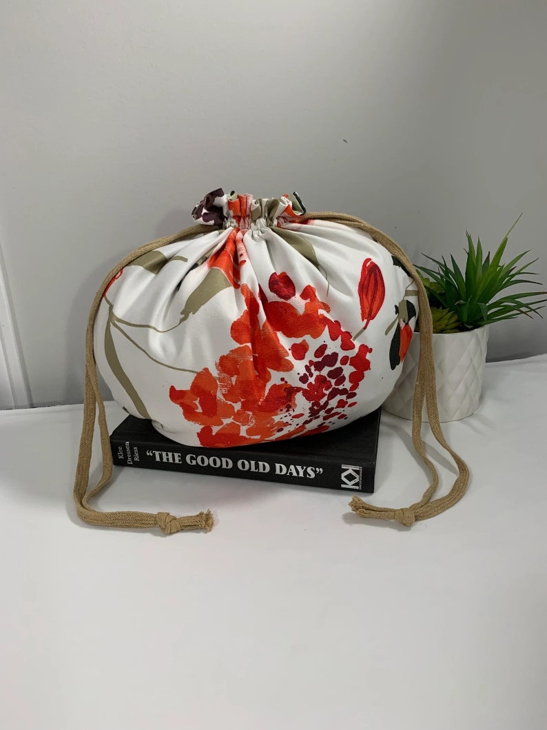 Puede incluir: Una bolsa de cord&oacute;n blanco con estampado floral en tonos naranja, rojo y marr&oacute;n. La bolsa est&aacute; encima de un libro titulado "The Good Old Days".