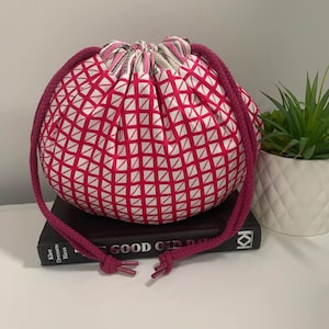 May include: A pink drawstring bag with a white geometric pattern. The bag is made of fabric and has a long, pink drawstring. The bag is sitting on top of a book.
