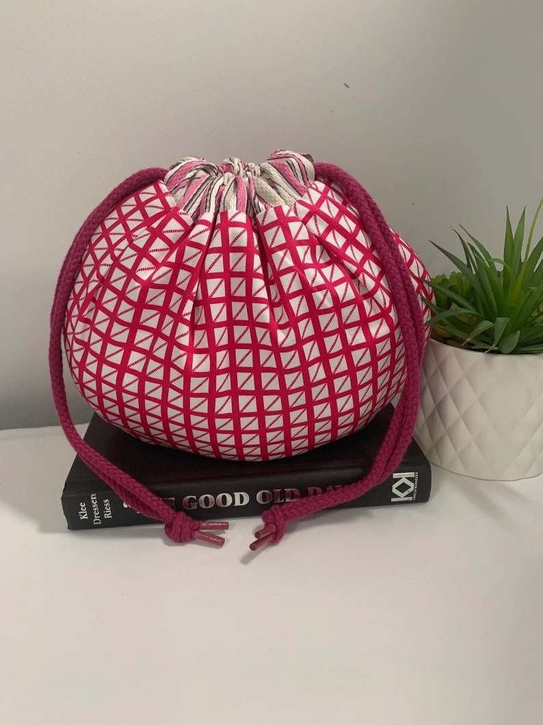 Puede incluir: Una bolsa de cord&oacute;n rosa con un patr&oacute;n geom&eacute;trico blanco. La bolsa est&aacute; hecha de tela y tiene un cord&oacute;n largo de color rosa. La bolsa est&aacute; encima de un libro.