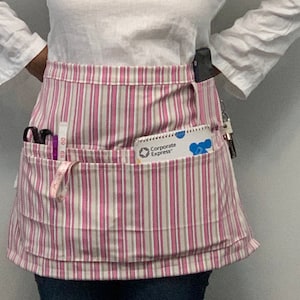 以下が含まれることがあります： ピンクと白のストライプの、ポケットが複数あるエプロン。エプロンには、メジャー、ハサミ、そして「Corporate Express」と書かれたメモ帳が入っています。