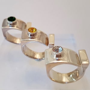 May include: Three silver rings with different coloured gemstones. The first ring has a green gemstone, the second ring has a yellow gemstone, and the third ring has a clear gemstone.