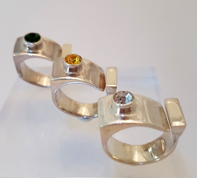 May include: Three silver rings with different coloured gemstones. The first ring has a green gemstone, the second ring has a yellow gemstone, and the third ring has a clear gemstone.