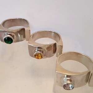 May include: Three silver rings featuring different coloured gemstones. Each ring has a rectangular band with a small gemstone set in the centre. The gemstones are green, yellow, and light blue.
