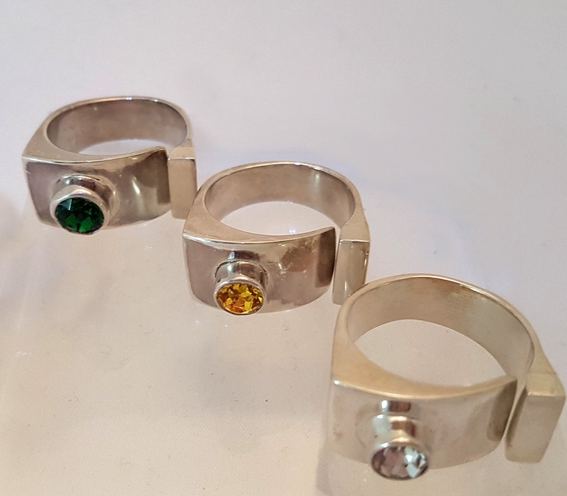 May include: Three silver rings featuring different coloured gemstones. Each ring has a rectangular band with a small gemstone set in the centre. The gemstones are green, yellow, and light blue.