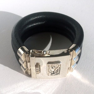 Pode incluir: Um bracelete de couro preto com um fecho prateado. O fecho tem um design decorativo e as letras "XXX" são visíveis.