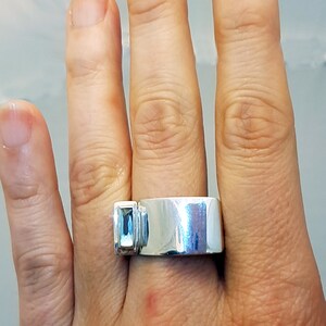 May include: A silver ring with a light blue gemstone set on the side. The ring is wide and has a smooth, polished finish.