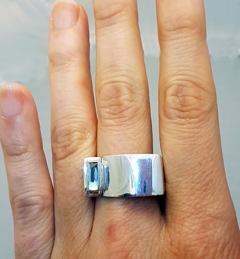 May include: A silver ring with a light blue gemstone set on the side. The ring is wide and has a smooth, polished finish.
