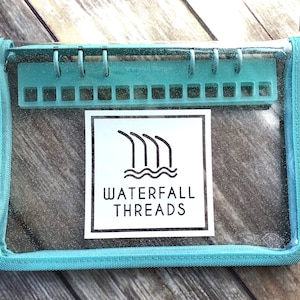 May include: A clear, rectangular pouch with a teal zipper and trim. Inside, a teal plastic organizer is visible. The pouch features a white label with the text "WATERFALL THREADS" and a logo of stylized waterfalls.