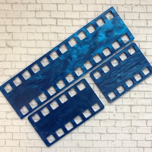 May include: Three blue film strip shaped wooden cutouts with a marbled effect. The cutouts are in varying sizes and are arranged diagonally against a white brick wall background. The cutouts have square openings.