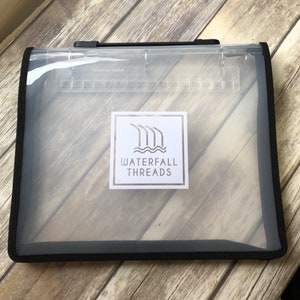 May include: Clear vinyl storage bag with a black trim and handle. The bag features a label with the text "WATERFALL THREADS" and a logo of stylized waterfalls. The bag is designed for organizing and storing items.
