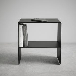 Puede incluir: Mesa auxiliar negra minimalista con una balda inferior y una superficie superior. Dos libros están en la balda lateral y un teléfono descansa en la parte superior. La mesa es de metal y tiene un diseño moderno.