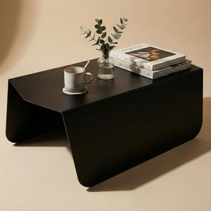 May include: A modern black coffee table with a minimalist design. The table has a rectangular top and curved legs. A mug, a small vase with greenery, and two books are on the table.