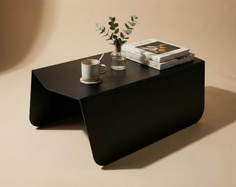 May include: A modern black coffee table with a minimalist design. The table has a rectangular top and curved legs. A mug, a small vase with greenery, and two books are on the table.