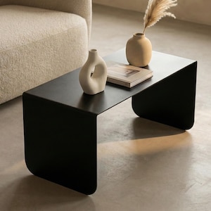 Könnte beinhalten: Ein moderner schwarzer Couchtisch mit minimalistischem Design. Der Tisch hat eine geschwungene, nahtlose Konstruktion. Auf dem Tisch stehen zwei Vasen, eine beige und eine weiße, und ein Buch. Der Tisch steht vor einem cremefarbenen Sofa.