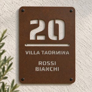 Könnte beinhalten: Ein rechteckiges Hausschild aus rostfarbenem Metall mit der ausgeschnittenen Zahl 20 oben, gefolgt vom Text VILLA TAORMINA und den Namen ROSSI BIANCHI darunter. Das Schild ist mit vier dunklen Schrauben an den Ecken befestigt.