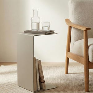Peut inclure: Une table d'appoint moderne de couleur neutre avec un rangement pour livres intégré. Une carafe en verre et un verre à boire sont posés sur le dessus de la table, avec un livre. Un fauteuil clair avec des accoudoirs en bois est à côté.