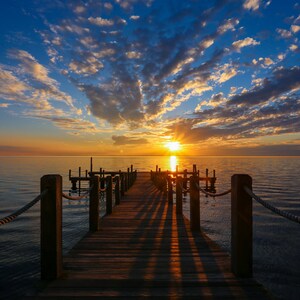 May include: A wooden dock extends out into the water towards a bright orange sunset. The sky is filled with fluffy white clouds. The water is calm and reflects the colors of the sunset.