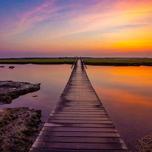 May include: A wooden walkway extends into a body of water at sunset. The water reflects the orange and pink hues of the sky.