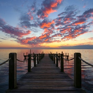 May include: A wooden pier extends into calm water under a colorful sunset sky. The sky is filled with orange and pink clouds, and the water reflects the colors. The pier has wooden posts and a rope railing.