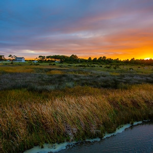 May include: A black and white striped lighthouse stands tall against a vibrant orange and blue sunset sky. The lighthouse is surrounded by a field of tall grass and a small body of water.