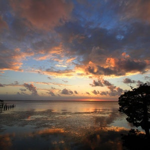 May include: A scenic sunset over a calm body of water with a wooden pier and a small gazebo in the foreground. The sky is a mix of pink, orange, and blue with fluffy clouds. The water reflects the colors of the sky.