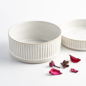 May include: Two white ceramic bowls with a vertical ribbed design. The bowls are empty and sitting on a white surface. There are several small pink flower petals and a brown flower bud on the surface.